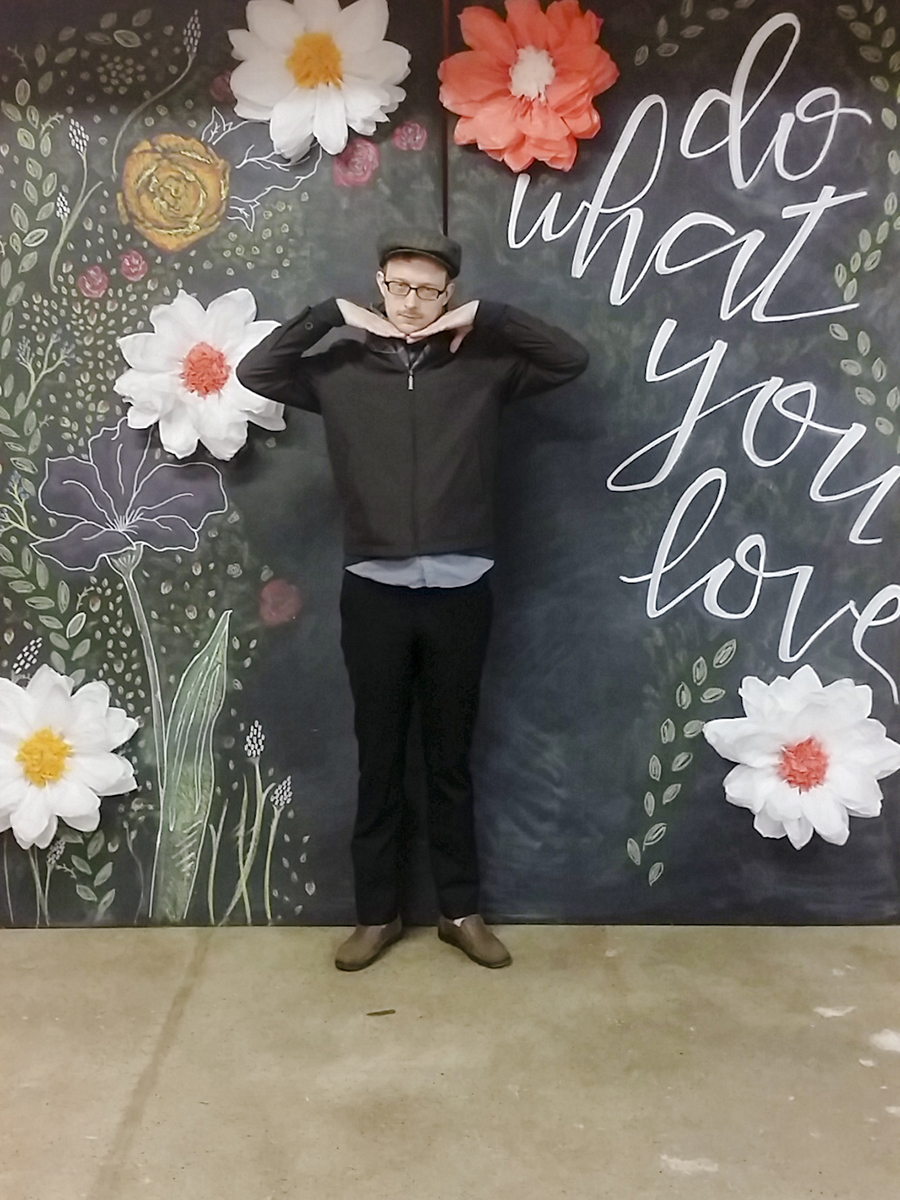 Ottie posing in front of blackboard 'Do What You Love' calligraphy and florals at Unglued craft fair.